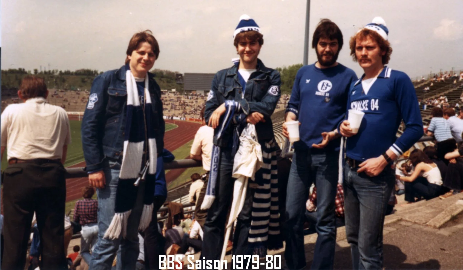 Vier Jugendliche stehen in einem Stadion, gekleidet in Fan- oder Mannschaftskleidung, mit den Schriftzügen "Schalke 04" und der Jahresangabe "1979-80" auf der Bildunterschrift, während sie Fans und Zuschauer im Hintergrund haben.