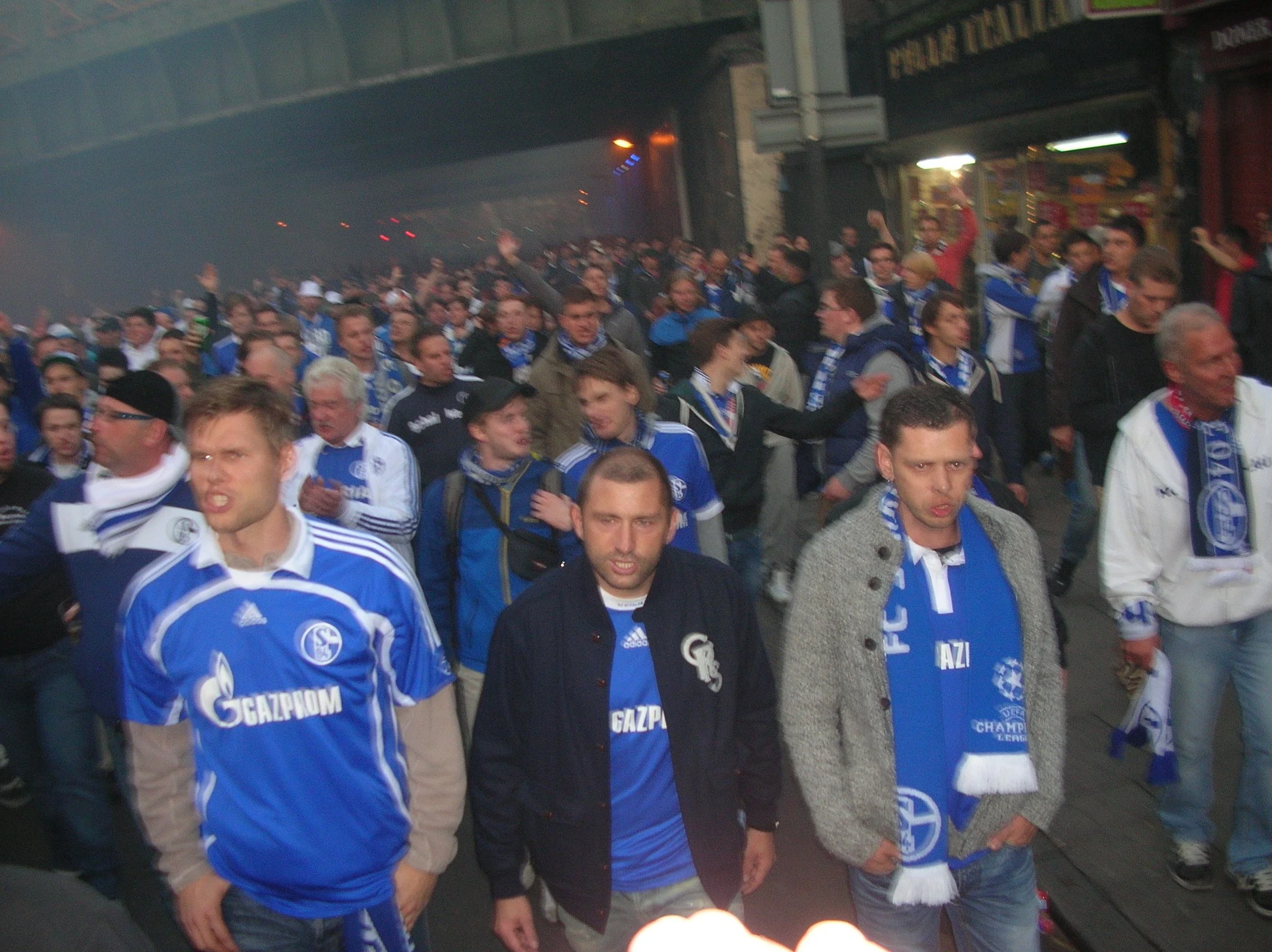 Menschenmenge in blauen Trikots, die auf eine Veranstaltung oder ein Ereignis warten, möglicherweise ein Fußballspiel, in einer städtischen Umgebung bei Nacht.