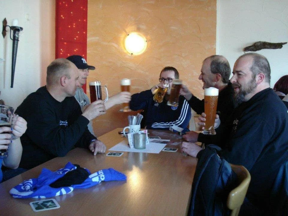Fünf Menschen sitzen an einem Tisch in einer Bar, lachen und heben ihre Gläser mit Bier zum Anstoßen.