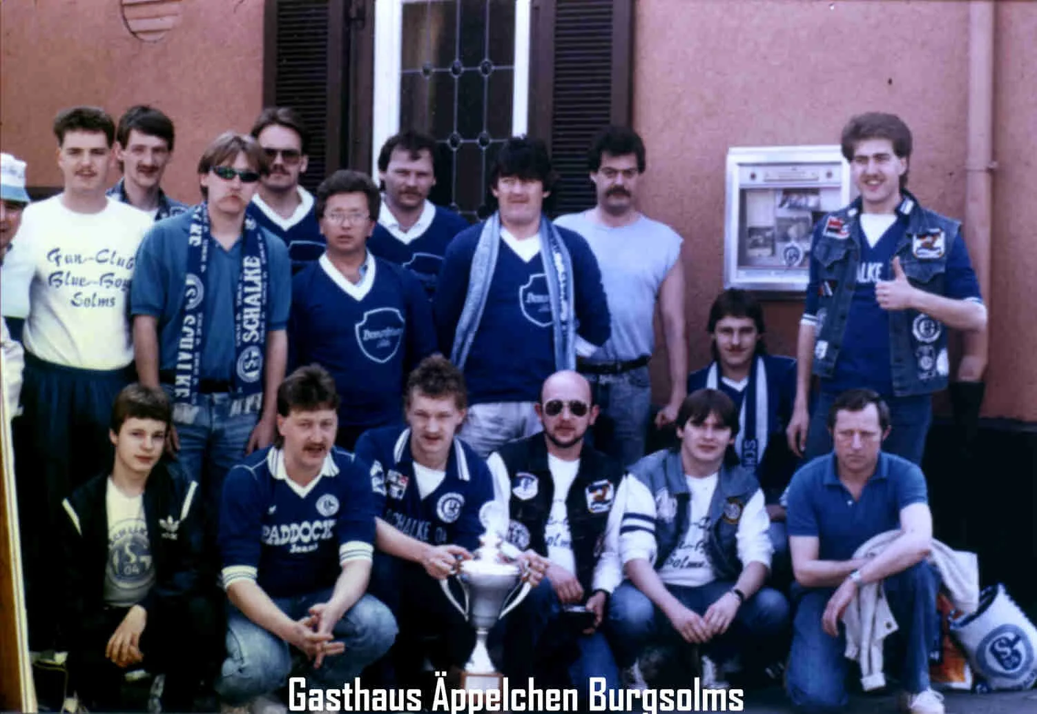 Gruppe junger Männer und Jungs, einige in Fan- oder Vereinskleidung, stehen und sitzen vor einem Mehrfamilienhaus, mit einem Pokal in der Mitte. Der Text unten lautet 'Gasthaus Äpplechen Burgsolms'.