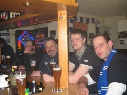Vier Männer sitzen an einer Bar, trinken Bier, und haben eine gesellige Zeit in einer Kneipe, mit Spielautomaten im Hintergrund.