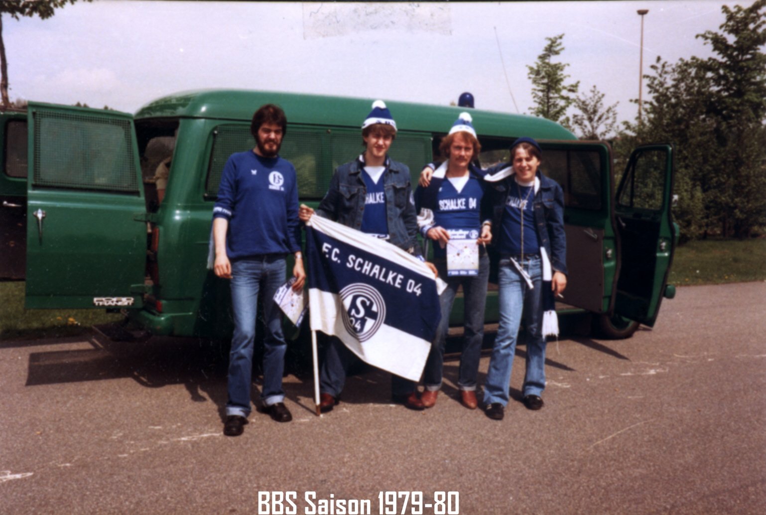 Vier Menschen stehen vor einem grünen Bus, tragen Schalke 04- der Kleidung und halten eine Schalke 04-flagge, wahrscheinlich bei einem Ausflug oder Spiel im Jahr 1979-80.