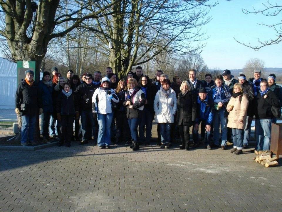 Gruppe von Menschen im Freien bei Sonnenlicht, einige tragen Winterkleidung, stehen vor Bäumen und einer grünen Infotafel auf gepflastertem Boden.