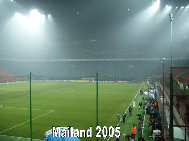 Leeres Fußballstadion bei Nacht mit Spielern und Personal am Spielfeldrand, keine Zuschauer auf den Rängen, Text 'Mailand 2005' im Bild.