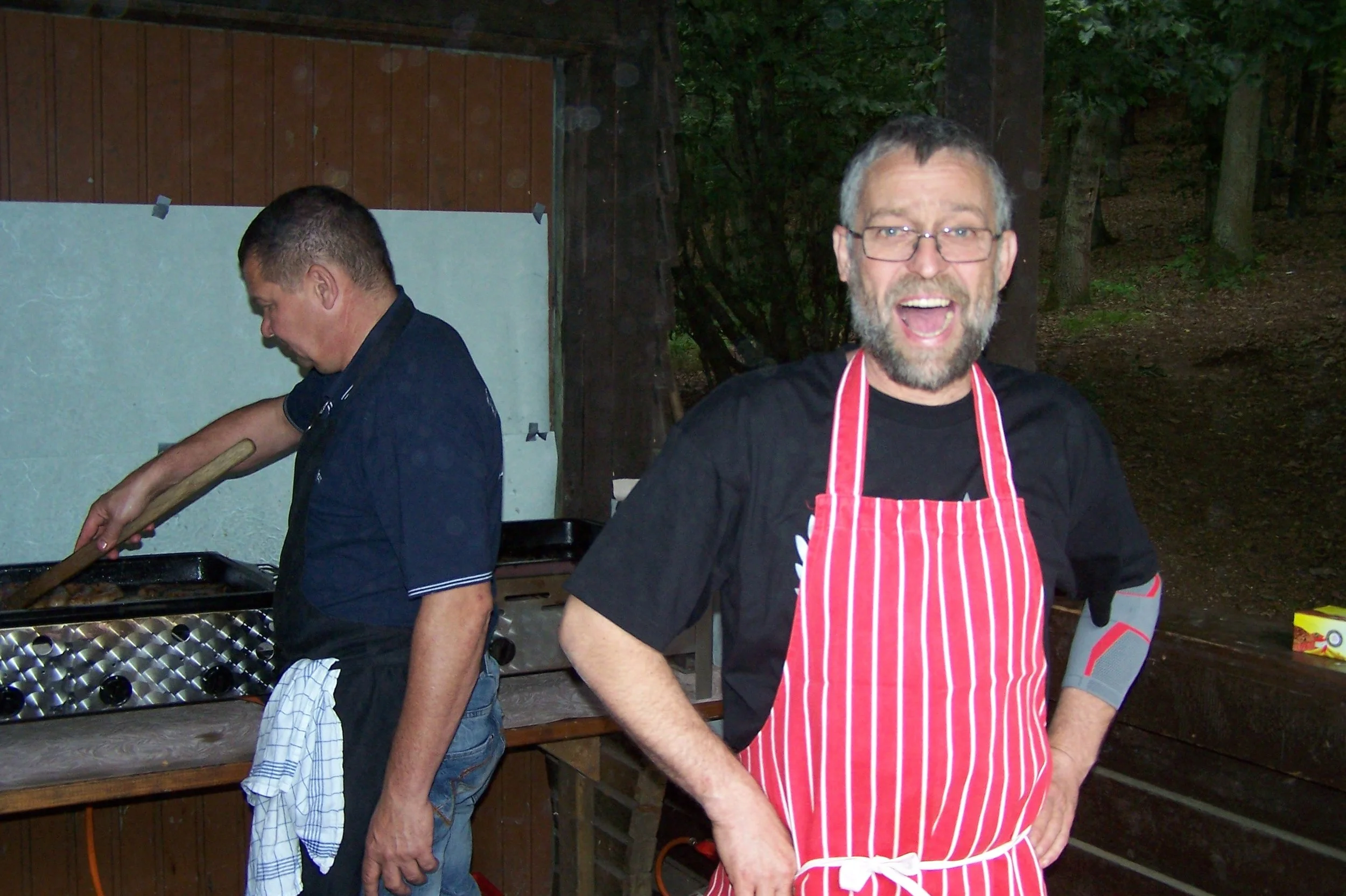 Zwei Männer beim Grillen im Freien, einer trägt eine rote und weiße Schürze, der andere steht neben dem Grill. Es ist dunkel und es gibt Bäume im Hintergrund.