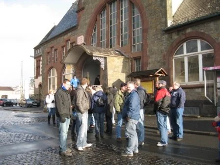 Eine Gruppe von Menschen vor einem alten Bahnhofsgebäude mit Steintreppen und großen Rundbögen