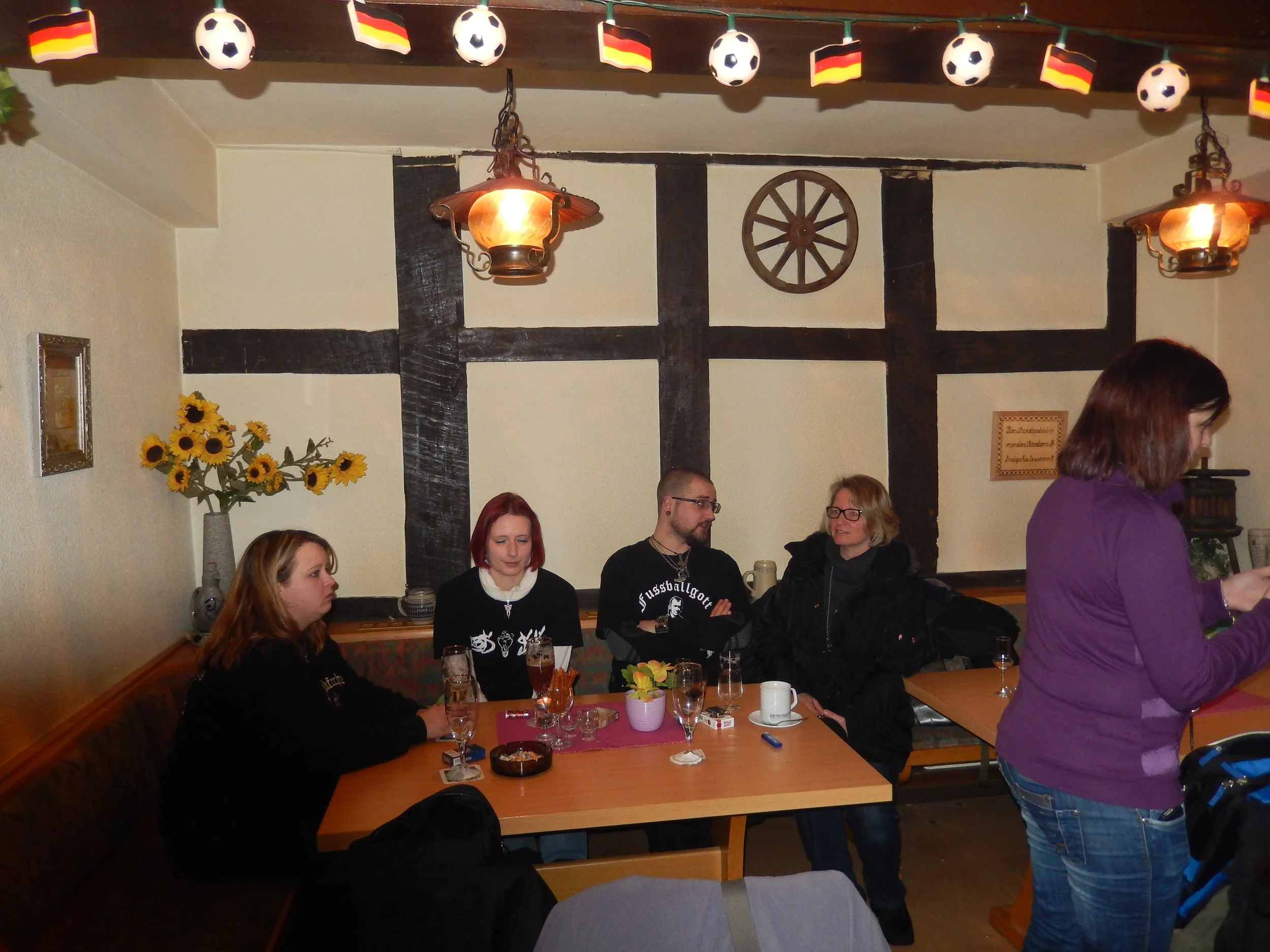 Menschen sitzen an einem Esstisch in einem gemütlichen Raum, der mit Wanddekorationen wie Sonnenblumen und Schwarz-Weiß-Elementen geschmückt ist. Es sind Getränke auf dem Tisch und eine Gruppe von Personen im Gespräch.