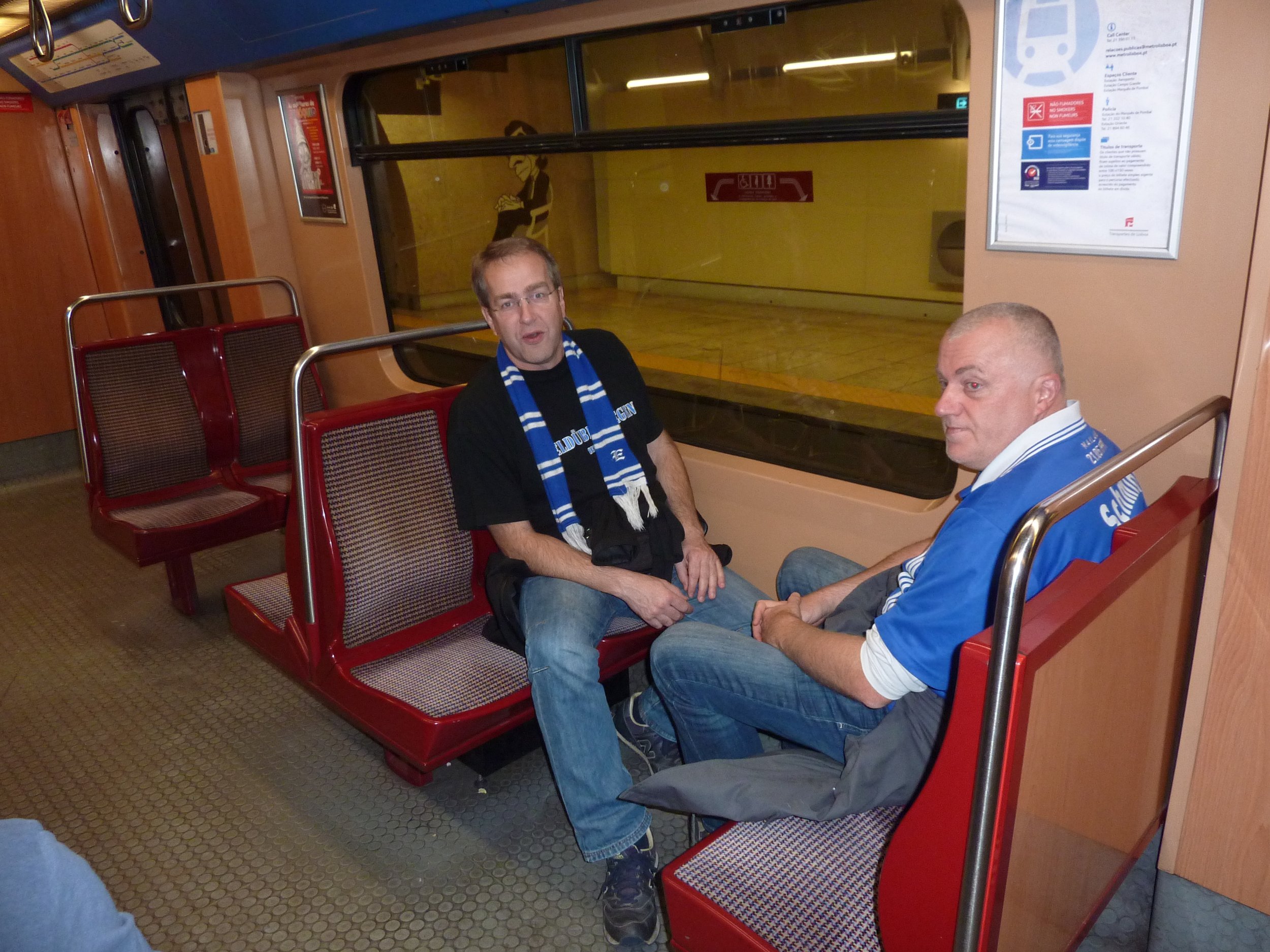 Zwei Männer sitzen auf einem roten Sitz im öffentlichen Nahverkehr. Der Mann auf der linken Seite trägt eine dunkel T-Shirt mit blauer Schal, der Mann auf der rechten Seite trägt eine blaue Jacke. Sie scheinen miteinander zu sprechen.