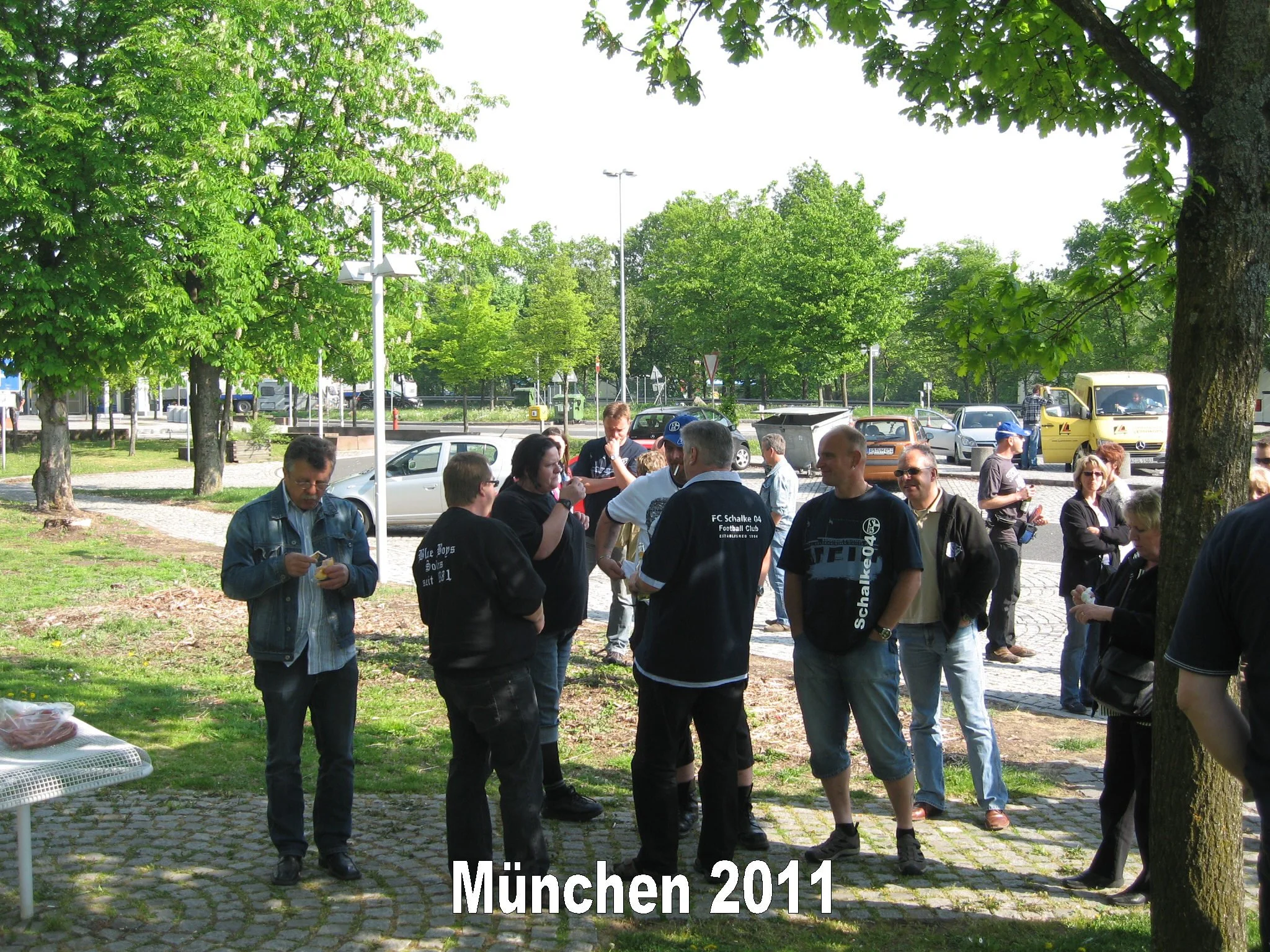 Gruppe von Menschen, die draußen an einem sonnigen Tag bei einer Veranstaltung in München 2011 zusammenstehen, umgeben von Bäumen und parkenden Autos.