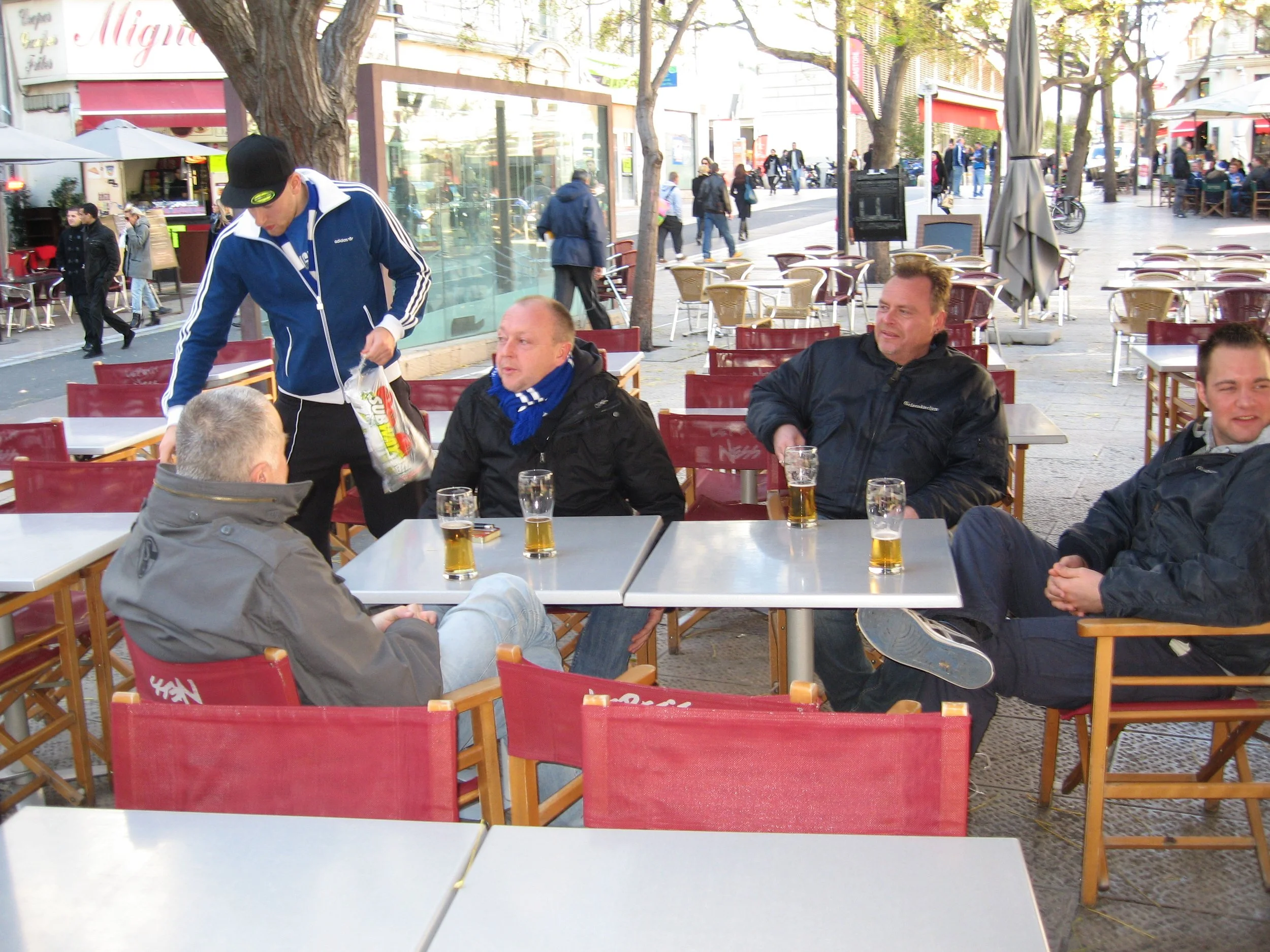 Menschen sitzen im Außenbereich eines Cafés und trinken Bier, während andere Spaziergänger die Straße entlanggehen, vorbei an Bäumen und Geschäften.