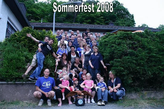 Gruppe von Menschen bei einem Sommerfest 2008, vor einem Haus mit grünem Busch und Bäumen im Hintergrund.