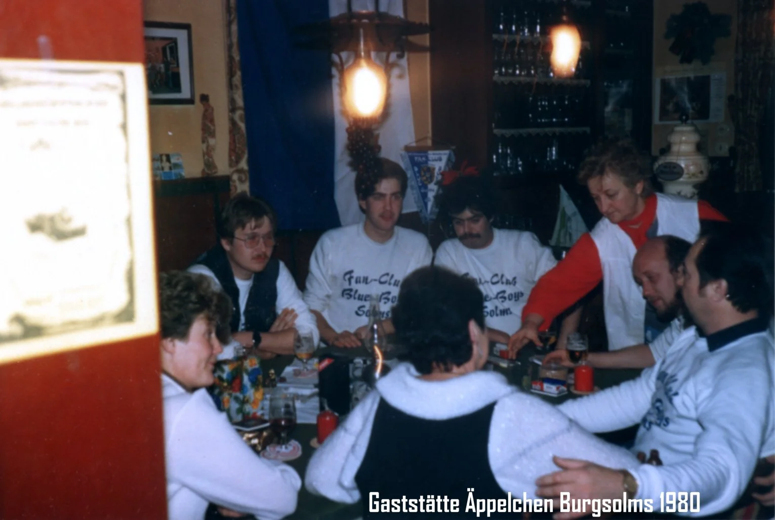 Eine Gruppe von Menschen sitzt um einen Tisch in einer Gaststätte, die im Jahr 1980 in Burgsolms namens "Äpplechen" aufgenommen wurde. Es gibt Getränke und Gegenstände auf dem Tisch, und eine Frau bedient die Gäste. Im Hintergrund sind Dekorationen u