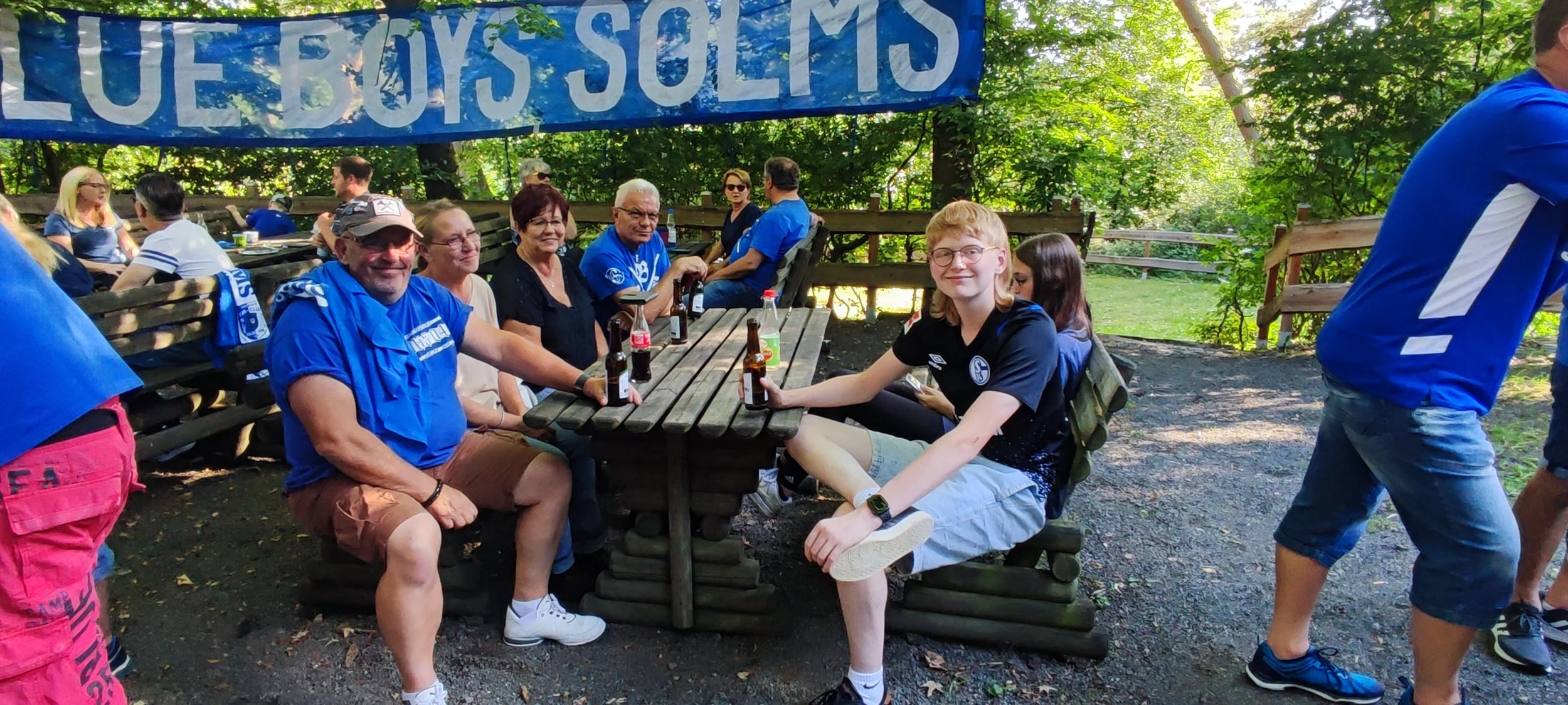 Gruppe von Menschen im Grünen, einige sitzen an einem Holztisch und trinken Bier, andere stehen oder sitzen im Hintergrund, alle tragen blaue Kleidung oder T-Shirts mit einem Logo, im Hintergrund ein blauer Banner mit weißen Buchstaben.
