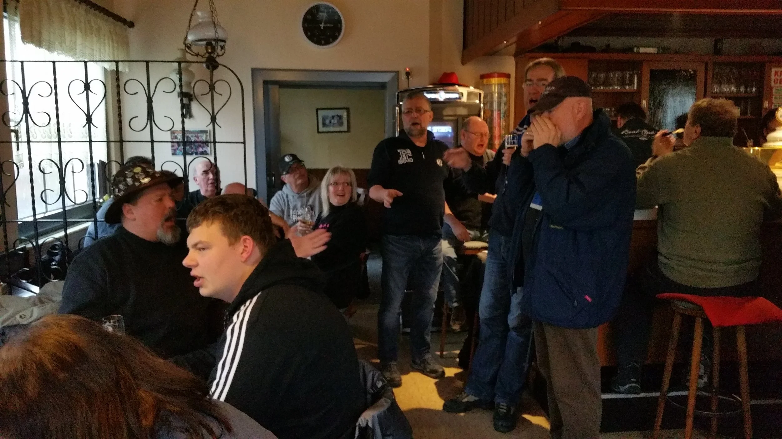 Mehrere Personen in einer Bar oder einem Bierlokal, einige sitzen an Tischen, andere stehen und unterhalten sich. Die Atmosphäre wirkt gesellig und lebhaft, mit dunkler Holzdekoration und Glasregalen im Hintergrund.