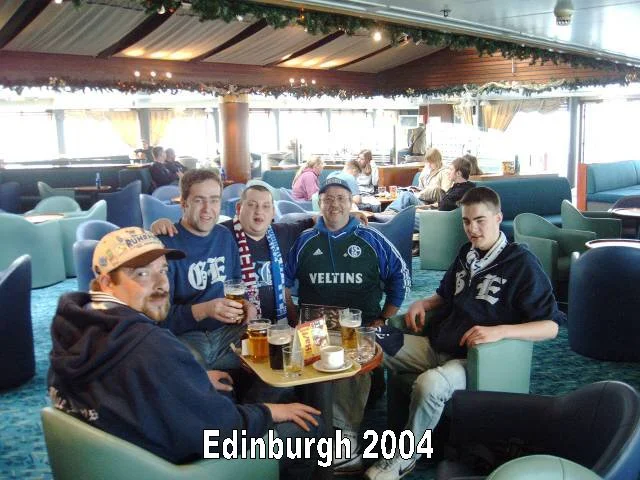 Fünf junge Männer sitzen in einer Bar oder einem Café in Edinburgh, 2004, trinken Bier und lachen, um einen kleinen Tisch mit Getränken. Im Hintergrund sind weitere Gäste und große Fenster mit Vorhängen.