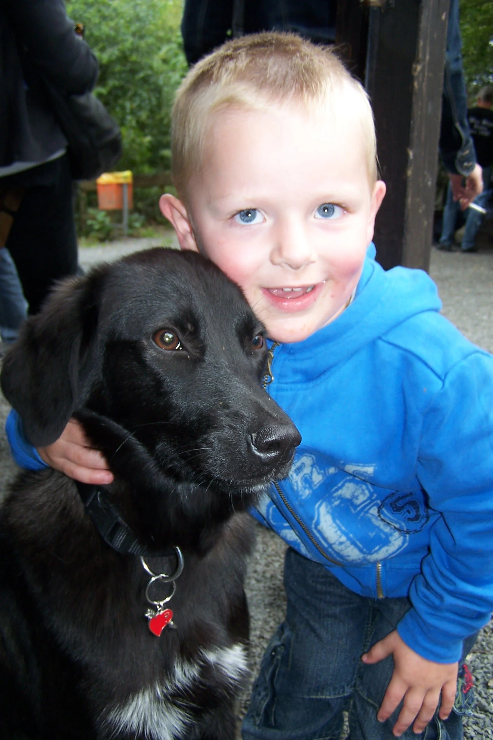 Ein Junge mit blonden Haaren und blauen Augen, der ein schwarzes Hundekind hält, im Freien mit Bäumen im Hintergrund.