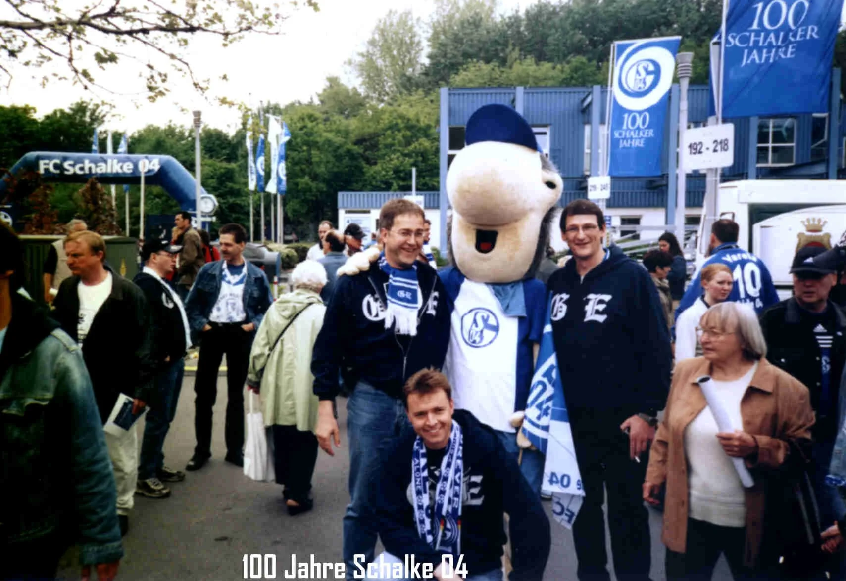 Menschen feiern das 100-jährige Jubiläum des Fußballclubs Schalke 04, einige tragen Schalke 04 Fanartikel, im Hintergrund Banner und Flaggen mit Schalke-Logo und Text."100 Jahre Schalke".