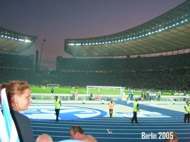 Fußballstadion in Berlin 2005 mit laufendem Spiel, Zuschauern im Hintergrund, mehrere Sicherheitskräfte auf dem Spielfeld, dunkler Himmel, Stadionlicht an.