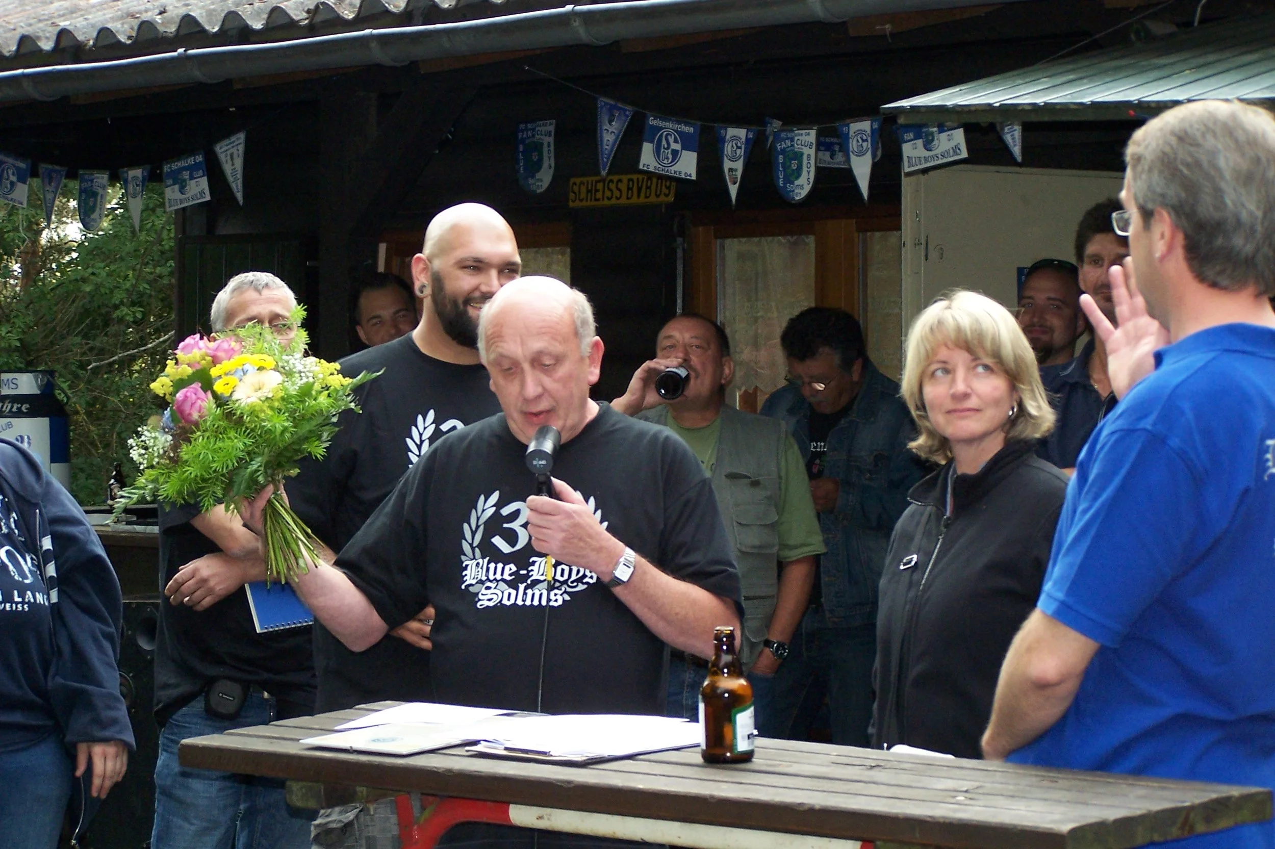 Menschen bei einer Feier, eine Person hält einen Blumenstrauß, im Hintergrund Fahnen eines Fußballvereins. Es scheint eine Zeremonie oder ein Fest zu sein.