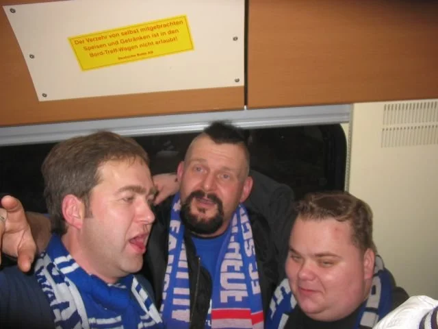 Drei Männer, die sich eng umarmen und freundlich in die Kamera schauen, tragen blauen und weißen Schal. Sie sitzen in einem öffentlichen Verkehrsmittel, vermutlich in einem Zug.