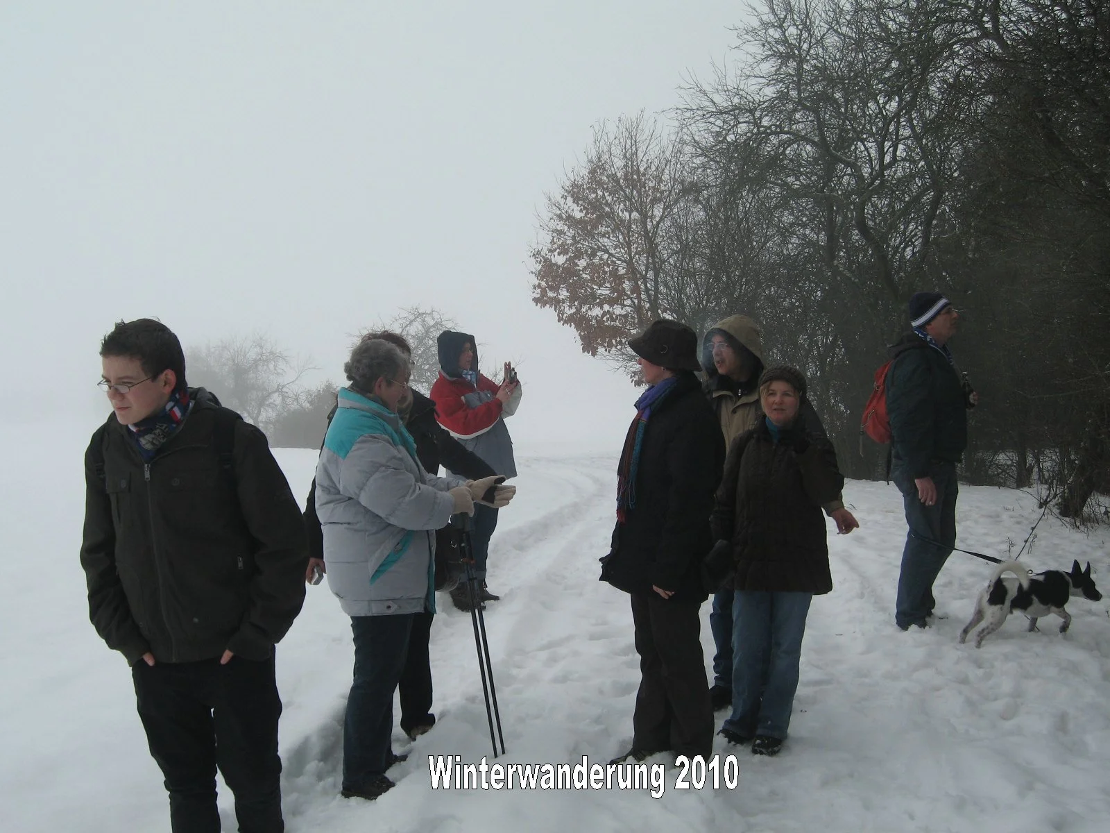 Gruppen von Menschen beim Winterwandern im Schnee, einige mit Kameras und einem Hund, im Hintergrund neblige Winterlandschaft mit Bäumen, Text "Winterwanderung 2010".