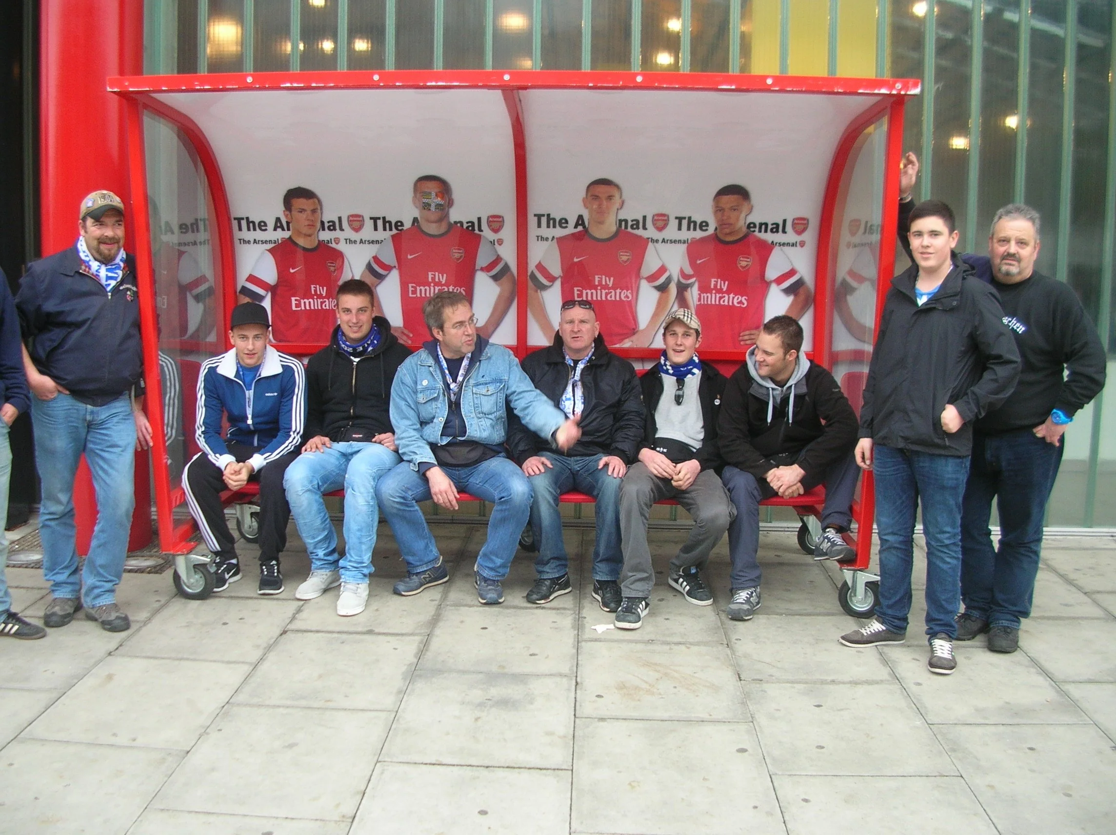 Gruppe von Menschen, einige sitzen und einige stehen vor einem roten Wartebereich mit Arsenal-Fußballspielern im Hintergrund, draußen vor einem modernen Gebäude.