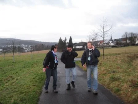 Drei Menschen gehen auf einem schmalen Weg in einer ländlichen Gegend, blicken auf den Boden und unterhalten sich. Der Himmel ist bewölkt, Bäume und Häuser sind im Hintergrund sichtbar.