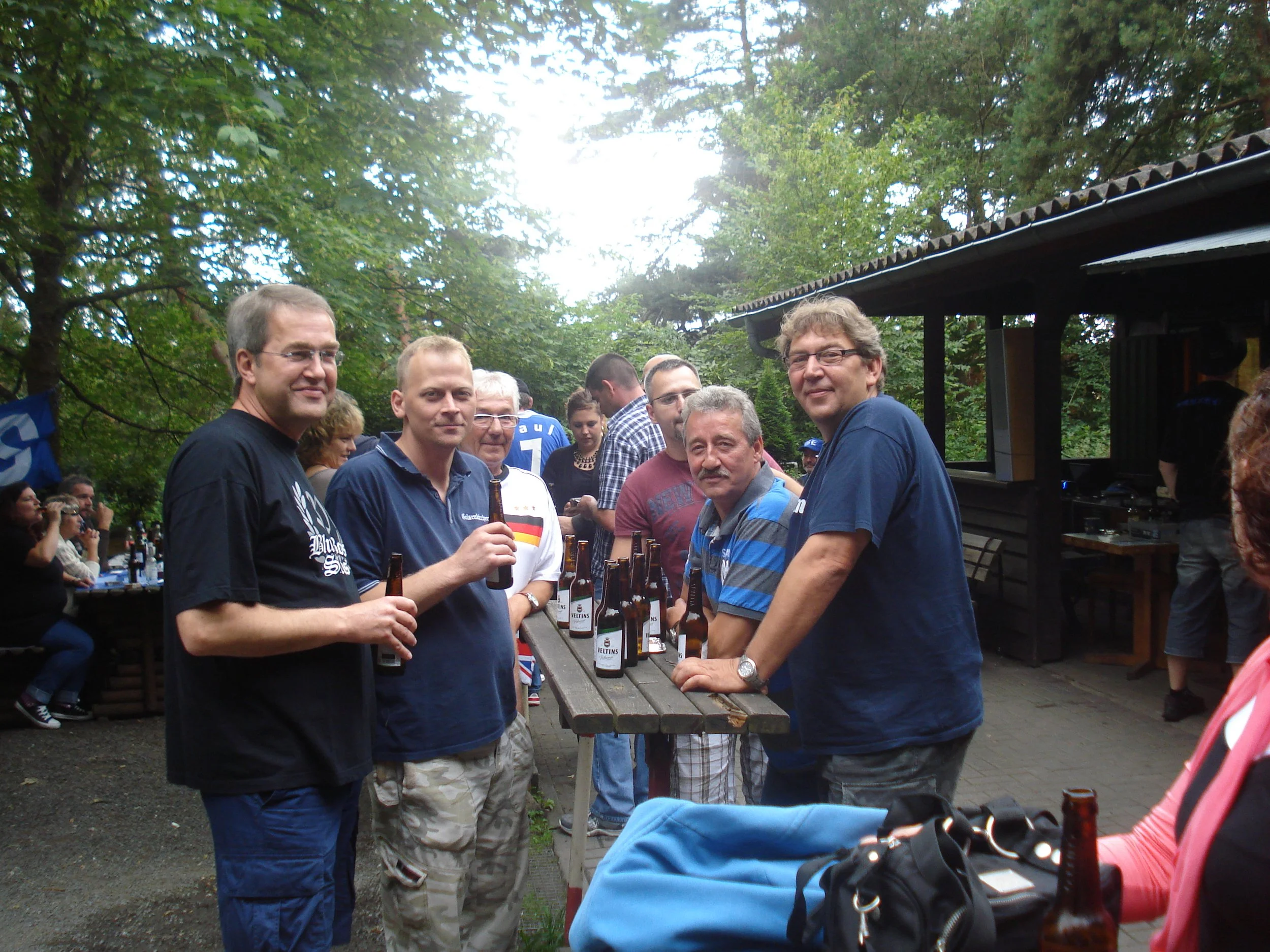 Eine Gruppe von Menschen steht um einen Holztisch mit Bierflaschen im Freien, umgeben von Bäumen und einem Holzgebäude bei einem gesellschaftlichen Treffen.
