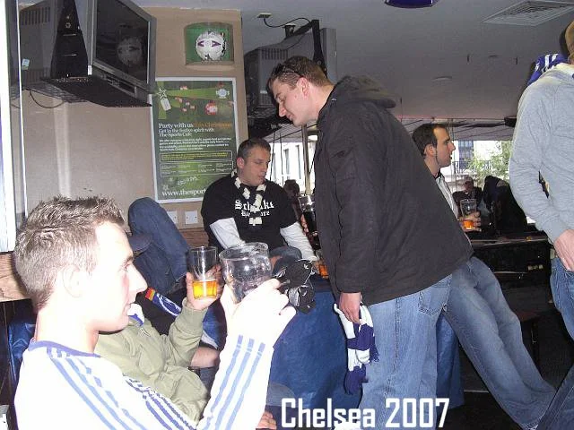 Menschen sitzen und stehen in einer Bar, einige trinken Bier. Es gibt einen Fernseher an der Wand, einen Fußball auf einem Regal und eine Tafel mit einem Spiel auf Deutsch.