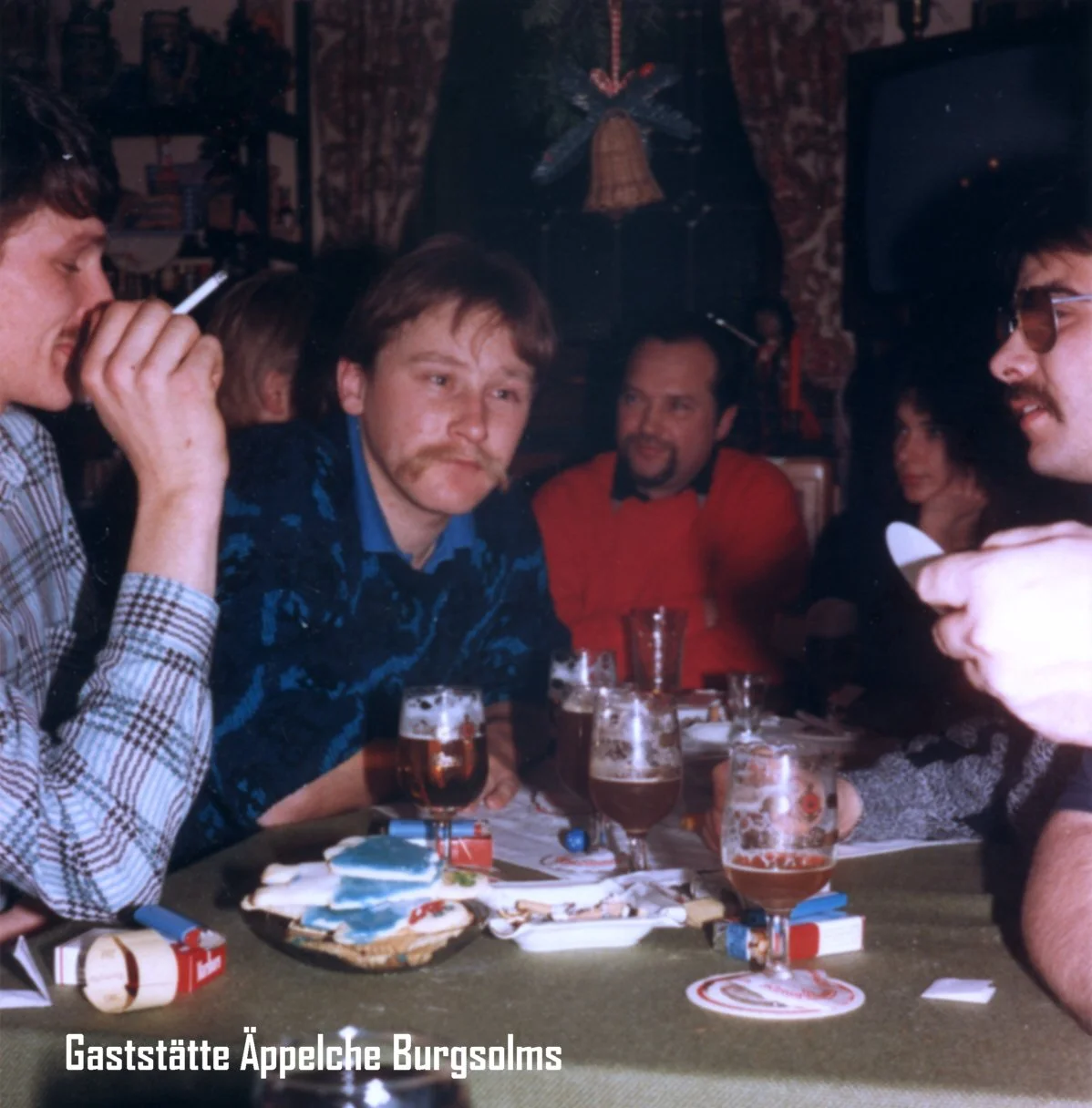 Eine Gruppe von Menschen sitzt um einen Tisch in einer gemütlichen Gaststätte, trinken Bier und essen Kekse, im Hintergrund Dekoration im typisch österreichischen Stil.