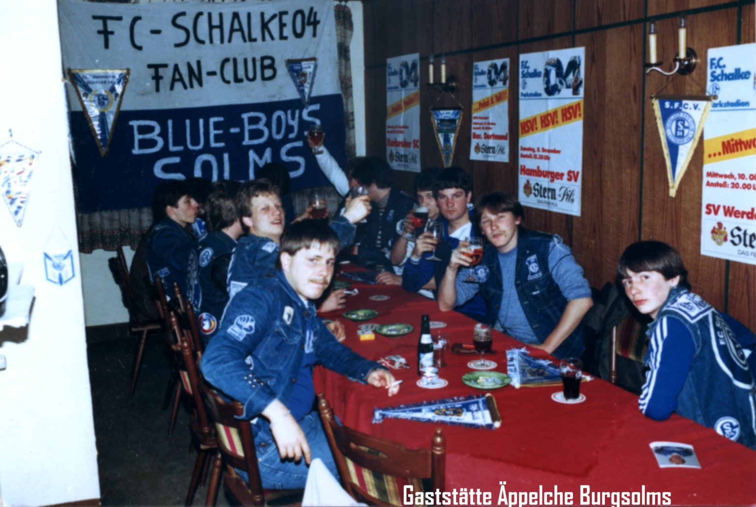Eine Gruppe junger Männer in Schalke 04 Fanoutfits sitzt an einem Tisch, der mit roten Tischtüchern bedeckt ist, in einem Raum mit Schalkeflaggen und Fanartikeln.