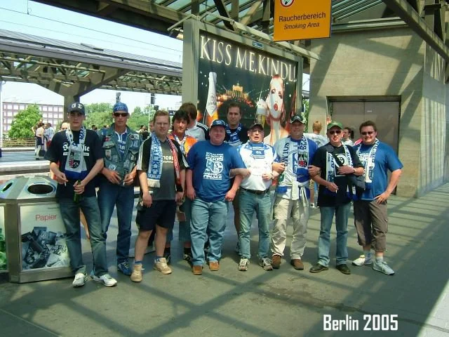 Eine Gruppe von Männern steht vor einem Eingangsbereich in Berlin im Jahr 2005, einige tragen blaue Schal und T-Shirts. Es gibt eine große Plakatwand im Hintergrund mit dem Text 'KISS ME KINDER', und ein gelber Bereich weist auf einen Raucherbereich 