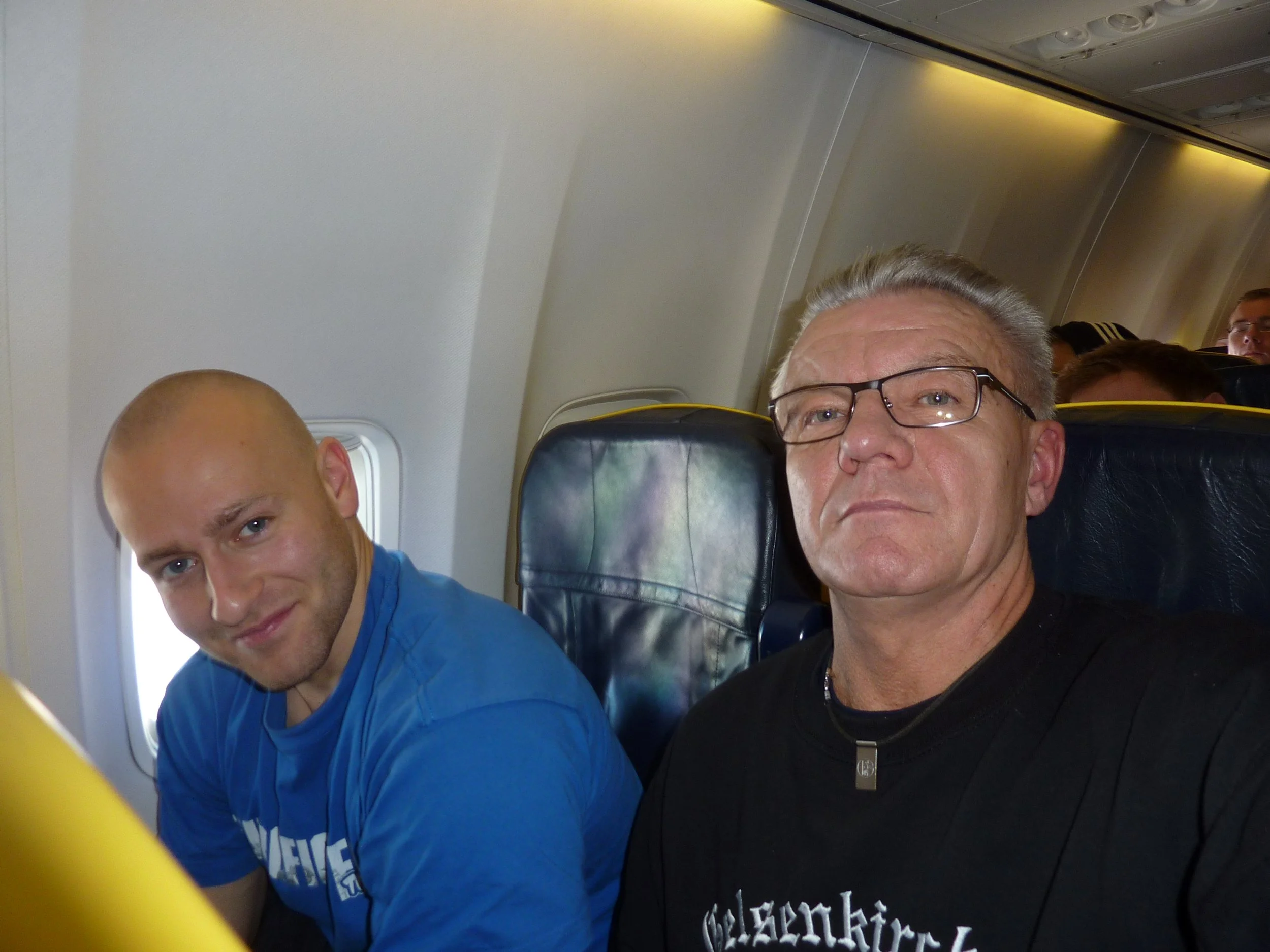 Zwei Männer sitzen in einem Flugzeug, einer trägt eine schwarze Jacke und Brille, der andere ein blauen Pullover.
