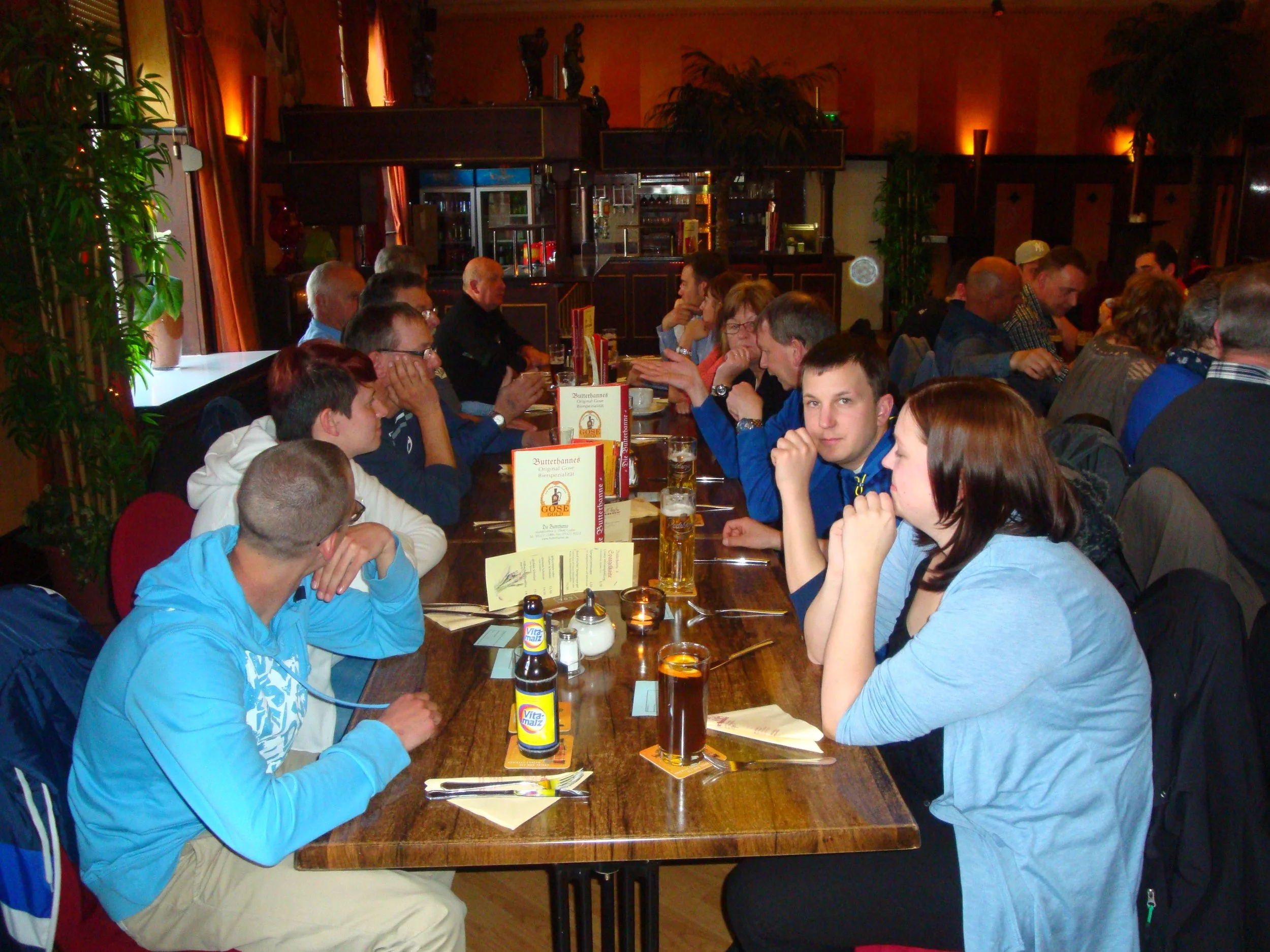 Mehrere Menschen sitzen an einem langen Tisch in einem Restaurant, trinken Bier und unterhalten sich. Im Hintergrund sind Pflanzen und Dekorationsartikel sichtbar.