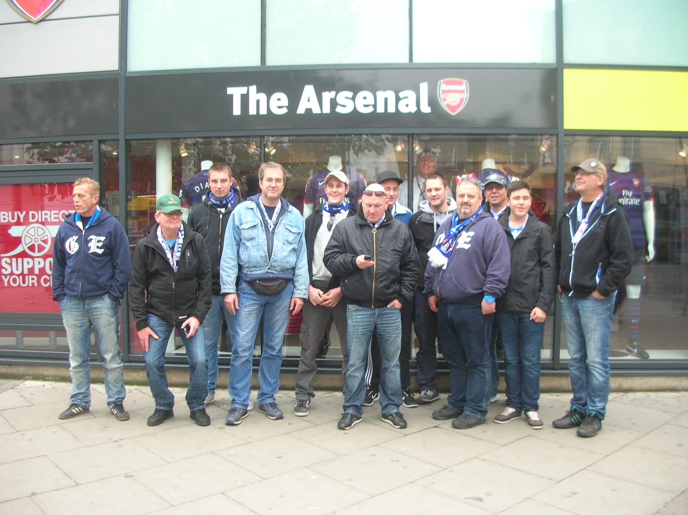 Eine Gruppe von Menschen steht vor einem Geschäft mit dem Schild 'The Arsenal' und ist wahrscheinlich Arsenal-Fans. Sie tragen Fanschals und Kleidung mit den Vereinsfarben.