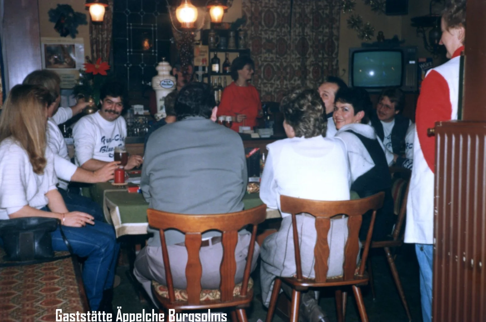 Menschen sitzen um einen Tisch in einer gemütlichen, dekorierten Gaststätte, im Hintergrund ein Fernseher und eine Bar. Eine Frau in rotem Oberteil steht hinter der Bar, während eine Frau in einem weißen Jacke mit rotem Rücken auf der rechten Seite s
