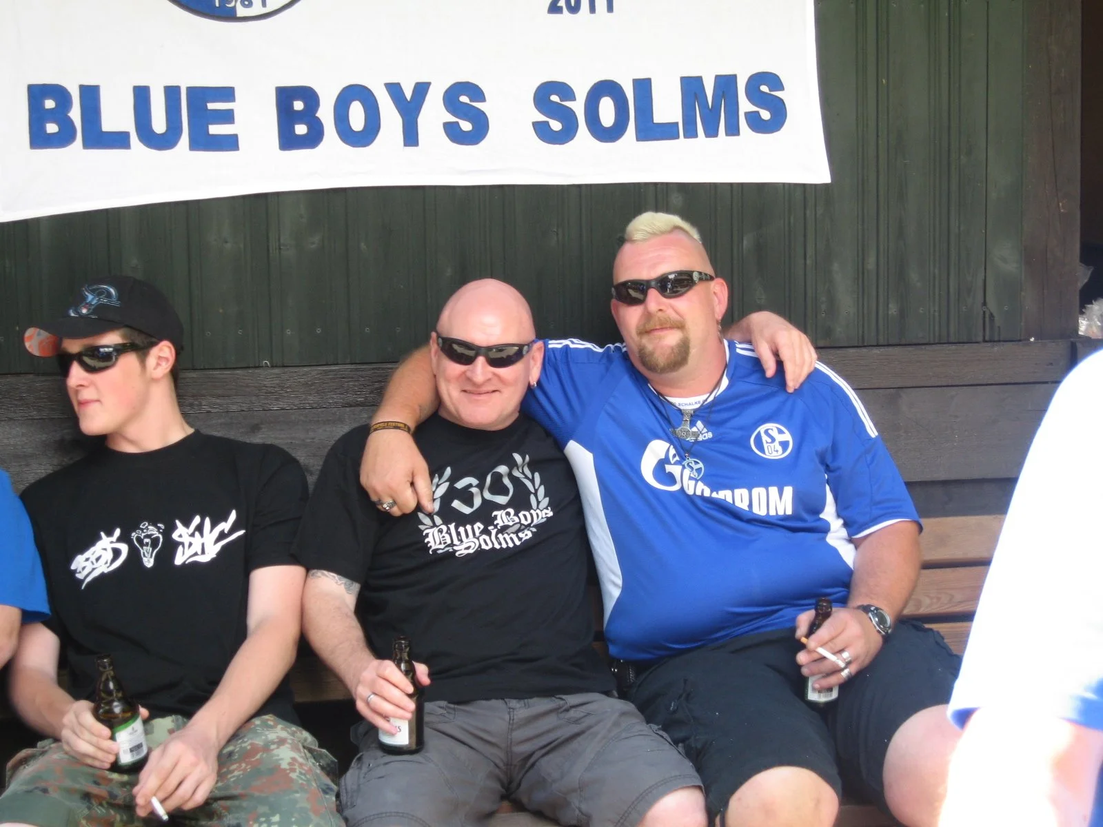 Drei Männer sitzen auf einer Bank, trinken Bier und posieren für ein Foto. Hinter ihnen hängt ein Banner mit der Aufschrift 'BLUE BOYS SOLMS'.