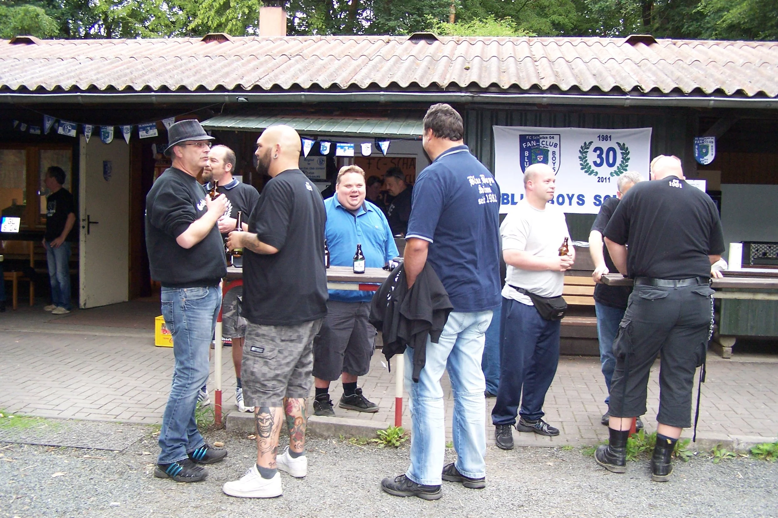 Eine Gruppe von Menschen und ein Kind, die auf einer Party im Freien am Stehtisch stehen. Es gibt eine große weiße Fahne mit blauer Schrift und die Jahreszahlen 1981 und 2011, sowie emsige Dekorationen mit blauen Fahnen.