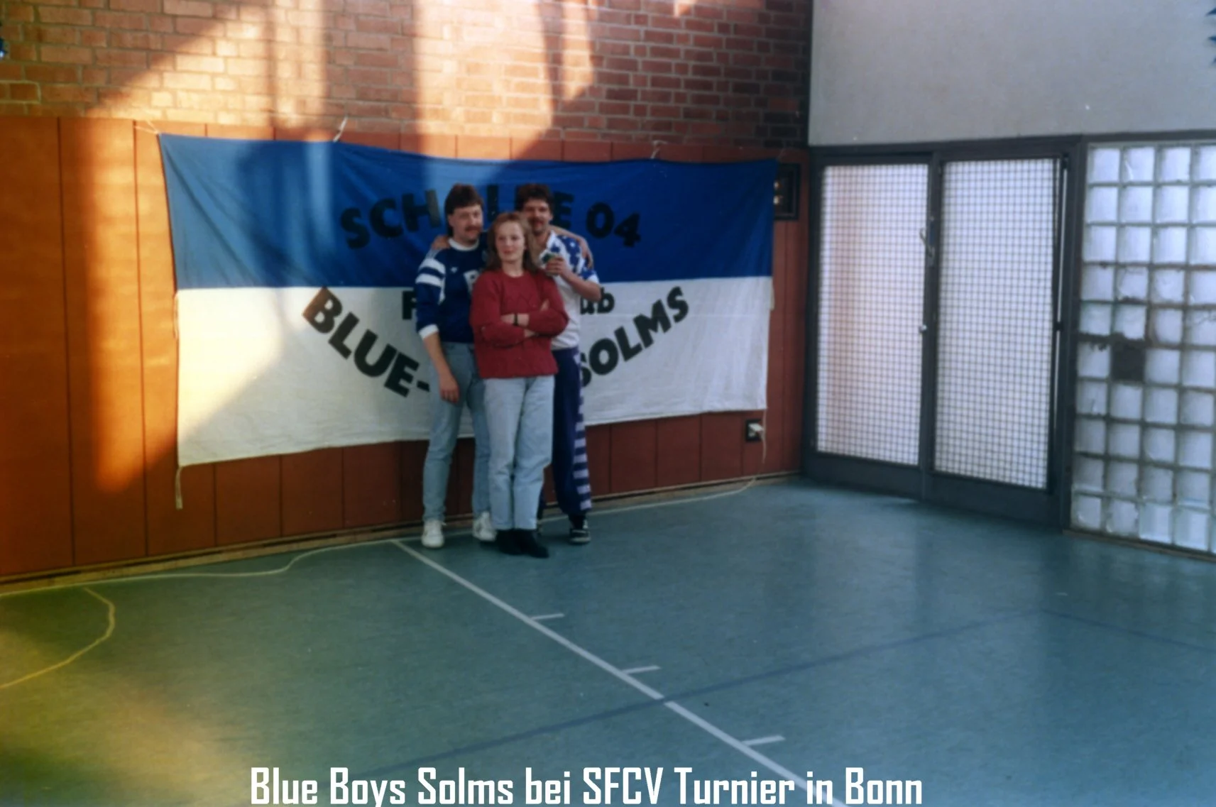 Drei Personen stehen vor einer blauen und weißen Fahne mit der Aufschrift 'Blue Boys Solms bei SFCV Turnier in Bonn' in einer Turnhalle.