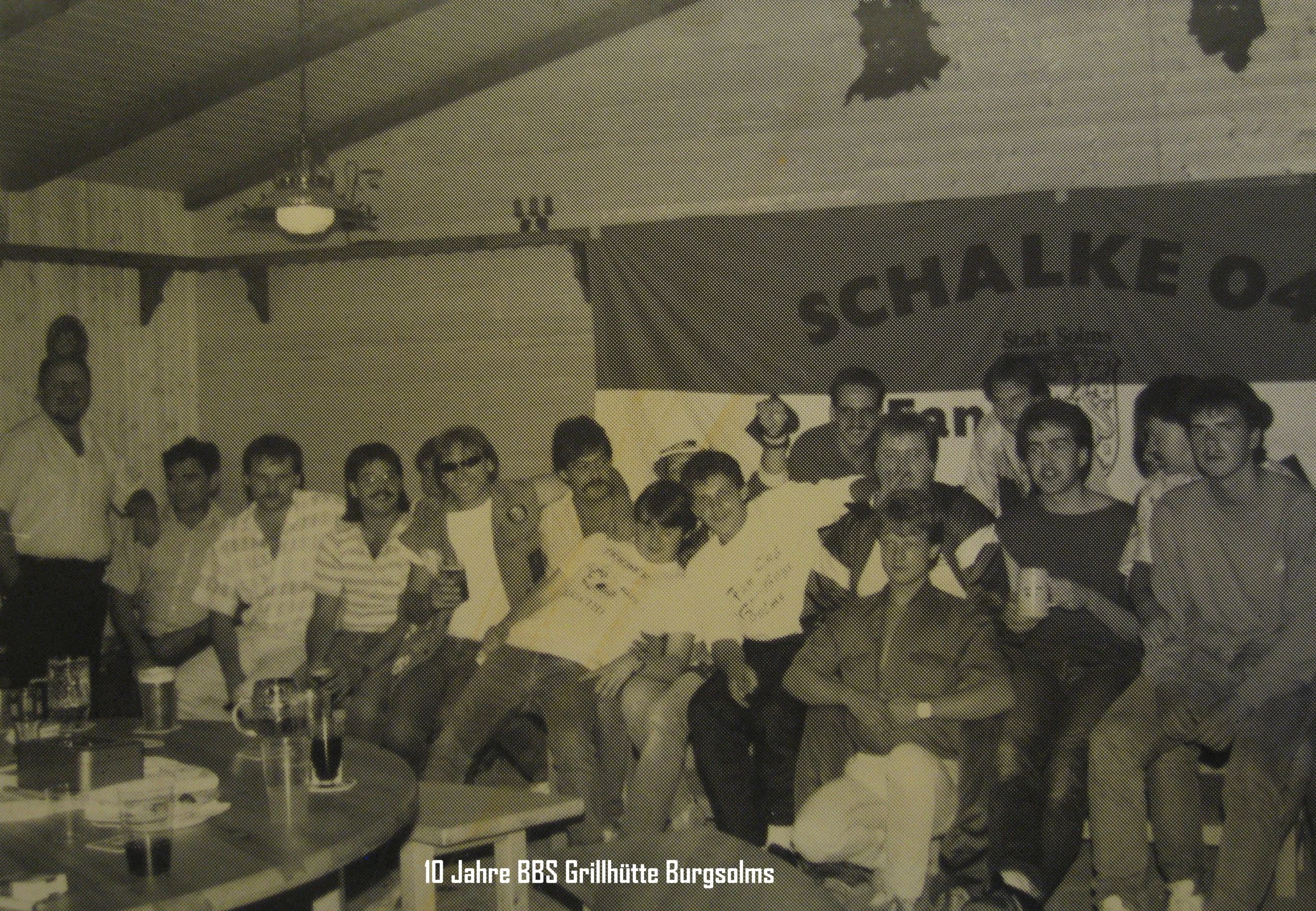 Ein schwarz-weiß-Foto von einer Gruppe von Menschen, die bei einem Jubiläum in einer Grillhütte zusammenstehen, mit einem Banner im Hintergrund, das den Anlass markiert, und mehreren Getränken auf dem Tisch. Der Text auf dem Bild lautet: "10 Jahre BB