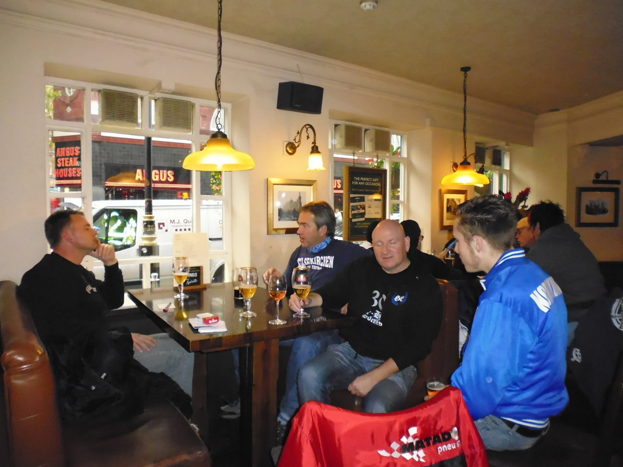 Mehrere Personen sitzen in einem Restaurant oder einer Bar, trinken Bier und unterhalten sich. Es gibt Fenster im Hintergrund mit Sicht auf die Straße, wo ein Geschäft namens "Angus Steakhouses" sichtbar ist.