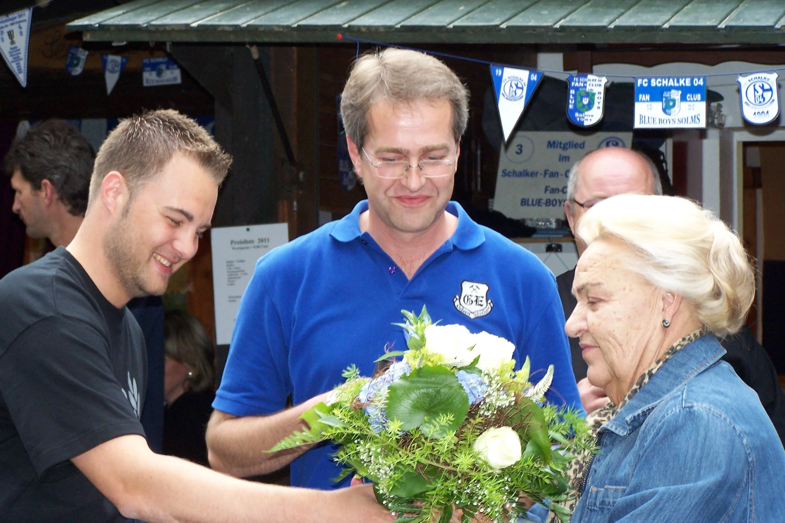Eine Gruppe von Menschen, darunter eine ältere Frau, empfangen eine junge Frau mit einem Blumenstrauß bei einer Feier. Sie lachen und umarmen sich in einem festlichen Umfeld mit Schleifen und Bannern im Hintergrund.