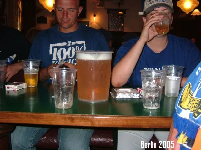 Zwei Männer sitzen an einem Tisch in einer Bar oder einem Restaurant. Der Mann auf der linken trägt ein blaues T-Shirt mit weißen Aufdruck, der Mann auf der rechten trägt eine blaue Jacke oder Pullover und trinkt aus einem Glas. Auf dem Tisch stehen 