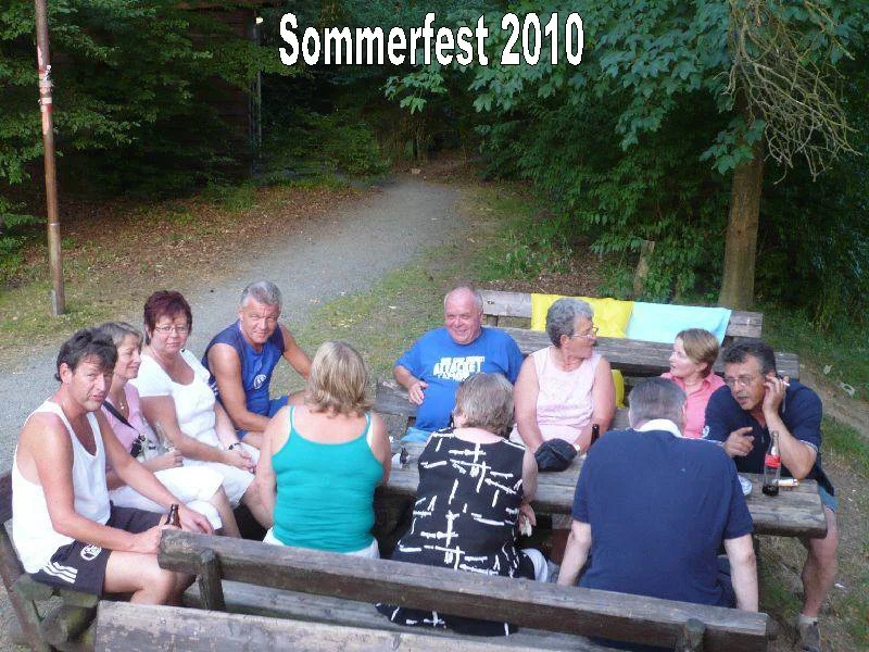 Gruppe von Menschen beim Sommerfest 2010 im Freien, die um einen Holztisch sitzen, mit Bäumen im Hintergrund.