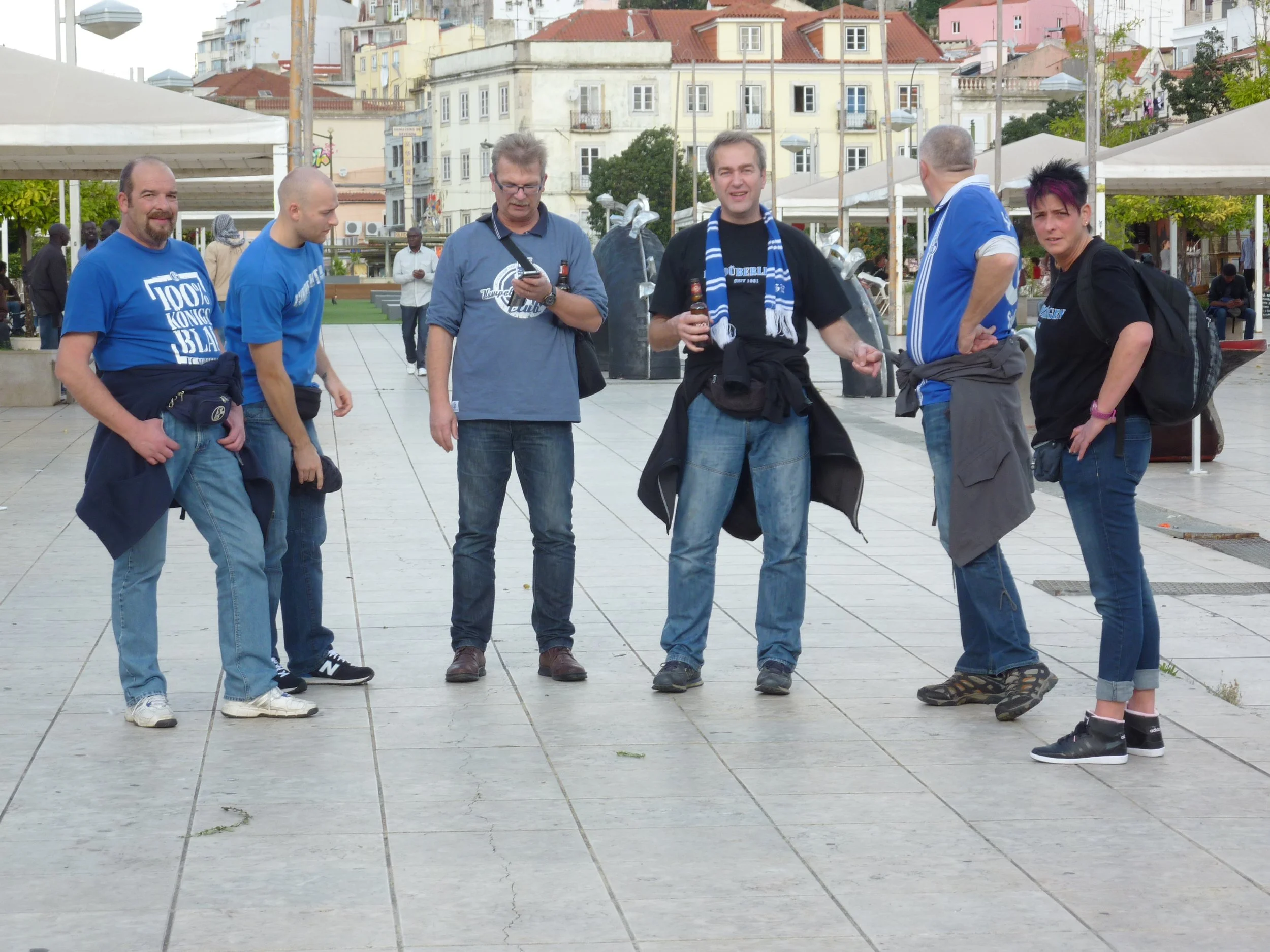 Sechs Menschen in einem öffentlichen Platz, einige tragen blaue T-Shirts mit weißen Aufdrucken, einige sprechen miteinander, eine Person liest eine Nachricht auf dem Handy. Im Hintergrund stehen Hütten und Gebäude, einige bunte und hohe Häuser.