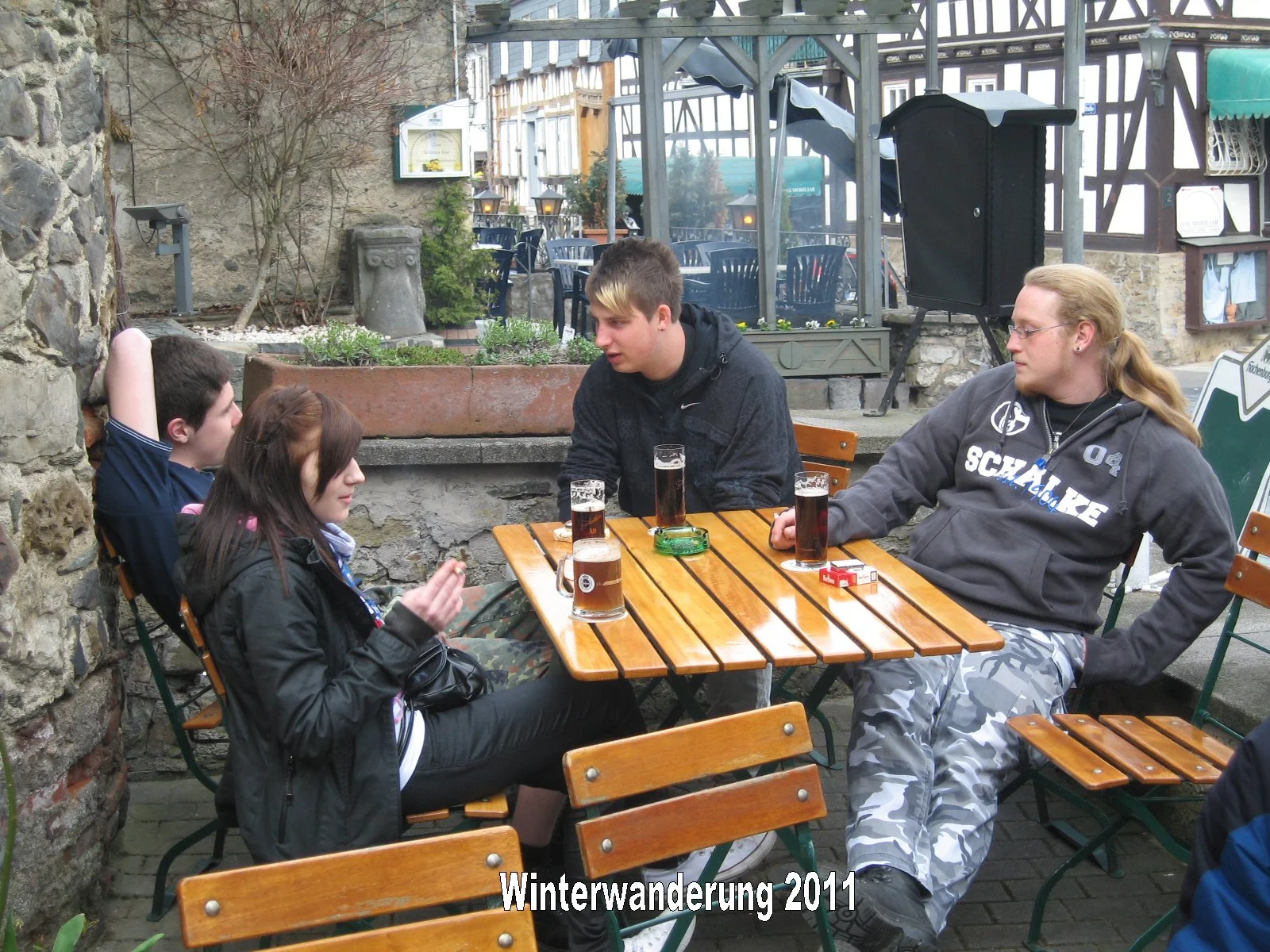 Eine Gruppe junger Menschen sitzt an einem Holztisch im Freien, trinkt Getränke und unterhält sich. Es ist offenbar kalt, da alle Jacken tragen. Der Hintergrund ist eine Mauer und eine Terrasse mit Tischen und Stühlen. Die Bildüberschrift lautet 'Win
