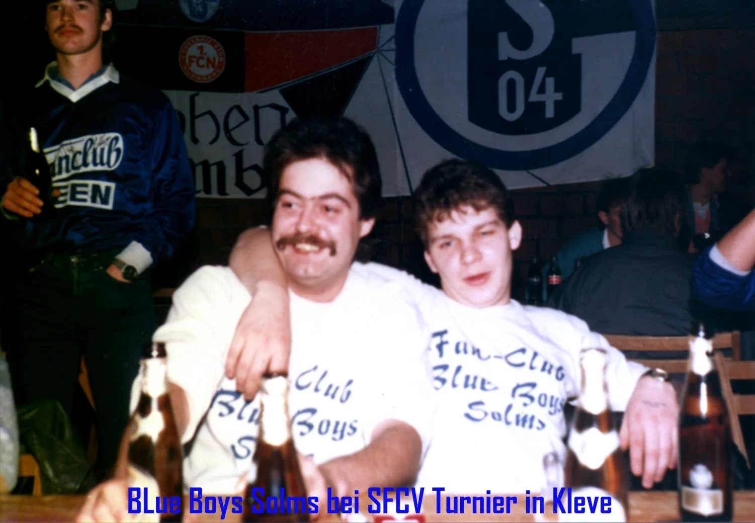 Zwei Männer mit weißen T-Shirts, auf denen 'Blue Boys Solms' steht, sitzen zusammen und lachen. Im Hintergrund sind weitere Menschen und ein Banner mit einem Fußballvereinssymbol. Es scheint eine Feier im Zusammenhang mit einem Fußballturnier in Klev