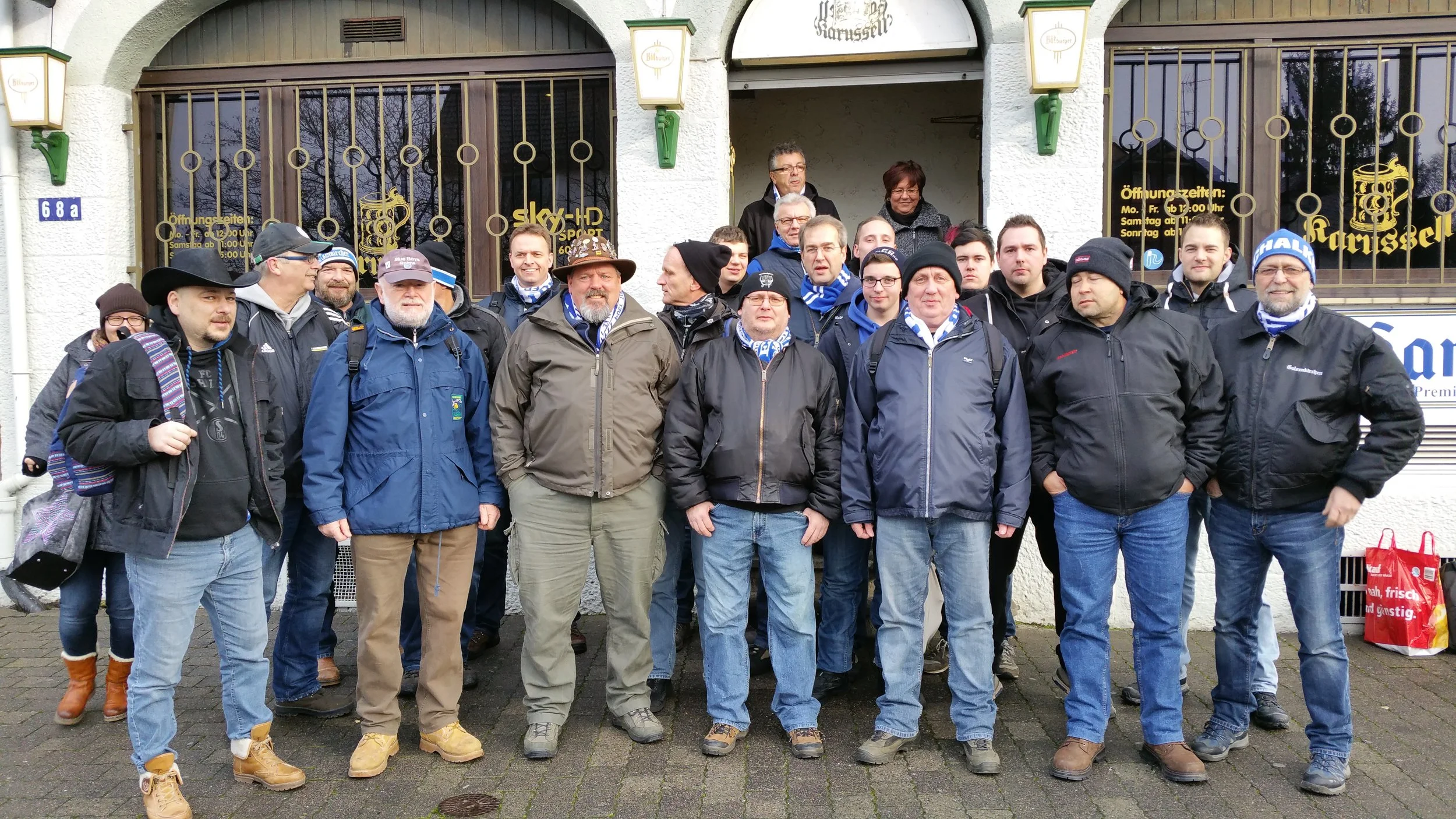 Gruppe von Menschen vor einem Gasthaus mit bayrischer Dekoration, einige tragen bayerische Trachten und Schals, vermutlich eine Fangruppe.