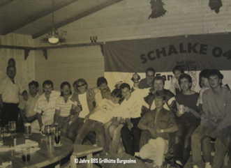 Jugendliche in einer Bar sitzen zusammen, einige stehen im Hintergrund, ein Schwarzwaldflagge mit 'Schalke 04' hängt an der Wand.