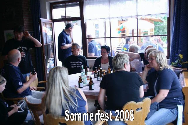 Menschen sitzen an Tischen in einer Gaststätte bei Sommerfest 2008, einige trinken und sprechen, im Hintergrund sind Fenster und Außenbereich sichtbar.